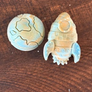 May include: Two molded soap pieces in blue and tan marbled colors. One piece is shaped like a spherical planet with raised surface continents, and the other piece is shaped like a rocket ship with a circular window and star details on the body.