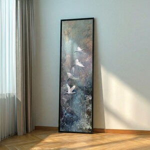 May include: A framed vertical art piece featuring a serene landscape with four white birds in flight against a backdrop of muted blues, greens, and browns. The artwork is encased in a black frame, adding a touch of elegance to the nature-inspired scene.