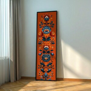 May include: A tall, rectangular framed wall art piece featuring a vertical, symmetrical floral pattern. The design displays stylized orange, blue, and dark teal flowers and leaves arranged in a repeating botanical motif against a solid orange background.
