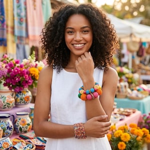 May include: A woman wearing a white dress and two colorful crochet bracelets. The bracelets feature a variety of bright colors, including pink, orange, blue, and green. The background includes flowers and market stalls.