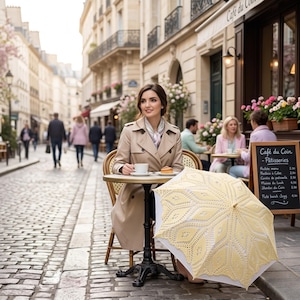 May include: A woman sits at a cafe table in Paris, France, wearing a tan trench coat and holding a cup of coffee. A yellow lace umbrella is open beside her. The cafe sign reads "Café du Coin - Pâtisseries."