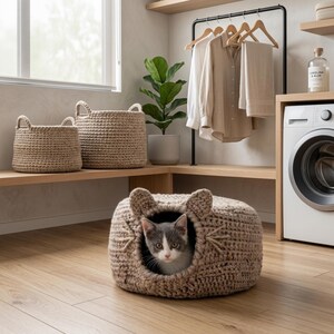 Peut inclure: Un chat gris et blanc regarde depuis un lit pour chat marron en crochet avec des détails en forme d'oreilles. Deux paniers tissés sont posés sur une étagère en bois, avec un troisième panier en arrière-plan. Une buanderie.
