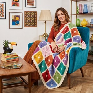 Könnte beinhalten: Eine bunte, handgehäkelte Decke mit einem geometrischen Muster aus quadratischen Feldern in verschiedenen Farben, darunter Rot, Gelb, Grün und Lila. Die Decke hat einen cremefarbenen Rand und ist über einem türkisfarbenen Sessel drapiert.
