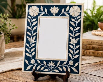 Op de afbeelding: Een rechthoekige fotolijst met een diep marineblauwe achtergrond en ingewikkelde witte bloemen- en rankinleg. De lijst wordt ondersteund door een donkerbruine houten standaard en toont een centraal wit rechthoekig vlak voor een foto.