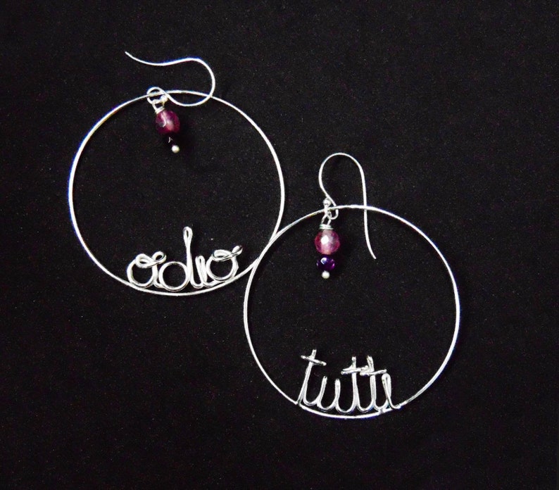 May include: A pair of silver hoop earrings with the words "solo" and "tutte" written in wire. Each earring has a dangling purple and pink bead.