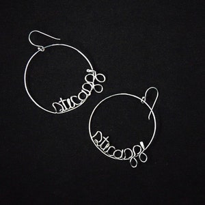May include: A pair of silver hoop earrings with the word "stuca" written in wire.
