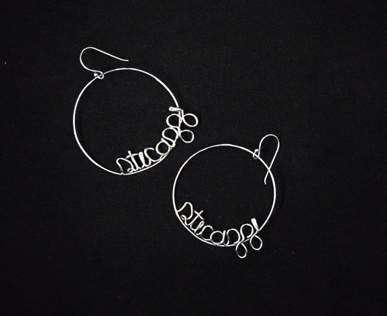 May include: A pair of silver hoop earrings with the word "stuca" written in wire.