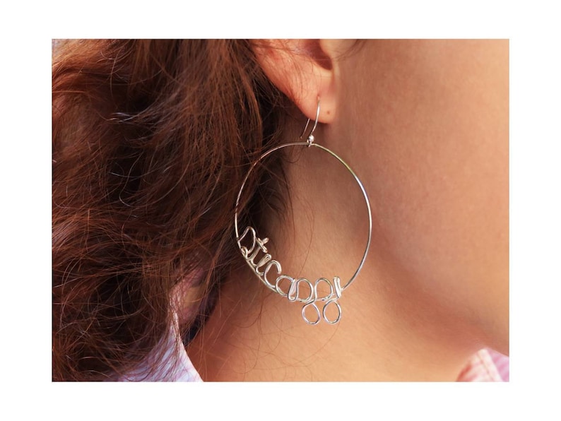 May include: Silver hoop earrings with the word "Chicago" spelled out in wire. The earrings are hanging from a person's ear.