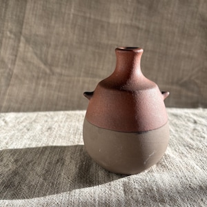 Könnte beinhalten: Eine kleine, handgefertigte Keramikvase mit zweifarbigem Design. Der obere Teil ist rotbraun, während die untere Hälfte dunkelbraun ist. Sie hat einen schmalen Hals, einen runden Körper und zwei kleine Griffe. Die Vase steht auf einer neutralfarbenen Leinenoberfläche.