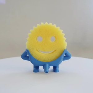 May include: A yellow sponge with a smiling face design, held by a blue plastic holder. The sponge has a sun-like shape with a series of points around the edge. The blue holder has arms and legs, giving the sponge a playful appearance. The product is designed for kitchen use.