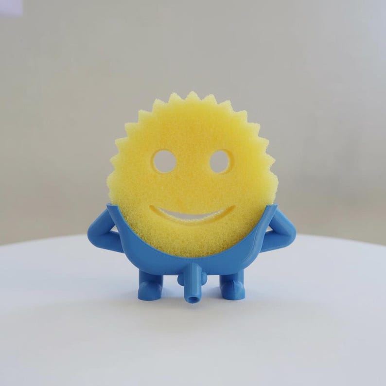 May include: A yellow sponge with a smiling face design, held by a blue plastic holder. The sponge has a sun-like shape with a series of points around the edge. The blue holder has arms and legs, giving the sponge a playful appearance. The product is designed for kitchen use.