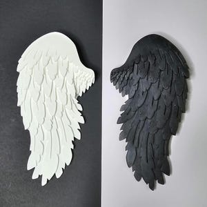 May include: Two decorative angel wings, one white and one black, with a feathery design. The wings are made of a textured material and are displayed against a black and white background.
