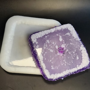 May include: A square, purple and white resin coaster with a white border and a glittery center. The coaster is sitting in a white silicone mold.