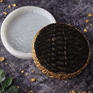 May include: A white, circular silicone mould with a textured interior is next to a dark brown, circular object with a gold-coloured, textured border. The dark brown object has a reptile skin pattern.
