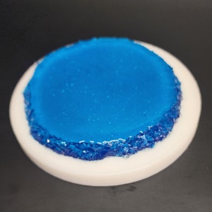 May include: A round, white coaster with a vibrant blue center. The edge of the blue center is textured with small, sparkling blue pieces. The coaster is set against a dark background.