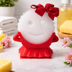 May include: A white sponge with a smiling face design, adorned with a red bow, and set in a red holder. The holder has a dress-like design. The image is set against a bathroom background.
