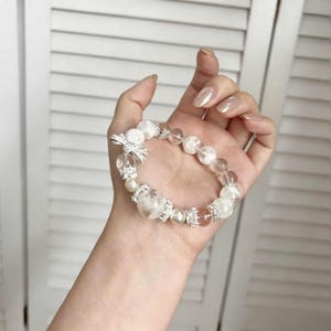 Könnte beinhalten: Ein filigranes Armband mit transparenten und weißen Perlen, verziert mit Perlen und glitzernden silberfarbenen Verzierungen. Das Armband hat einen charmanten Bogenanhänger und ist für eine elegante Ästhetik entworfen.