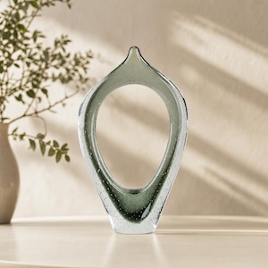 Op de afbeelding: Een traanvormige glazen sculptuur met een doorschijnende saliegroene tint en een heldere glazen rand. De binnenkant van het glas bevat talloze kleine, zwevende luchtbelletjes door de hele structuur, wat zorgt voor een getextureerd uiterlijk.