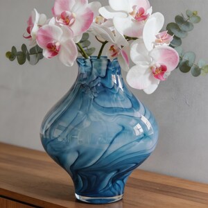 May include: A blue glass vase with a swirling pattern holds white and pink orchids and green eucalyptus leaves. The vase has a bulbous shape and sits on a wooden surface, creating a decorative floral arrangement.