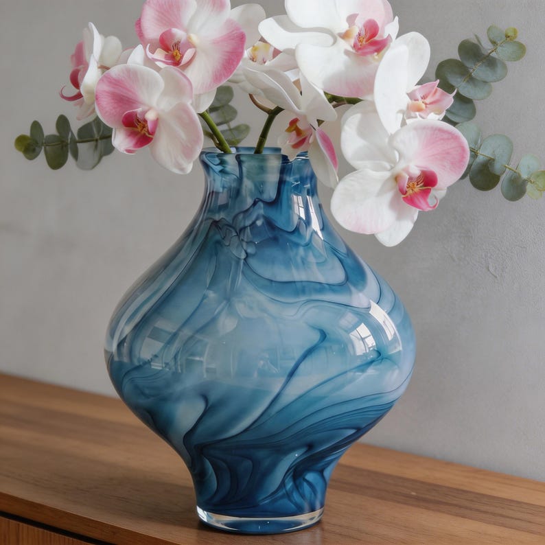 May include: A blue glass vase with a swirling pattern holds white and pink orchids and green eucalyptus leaves. The vase has a bulbous shape and sits on a wooden surface, creating a decorative floral arrangement.