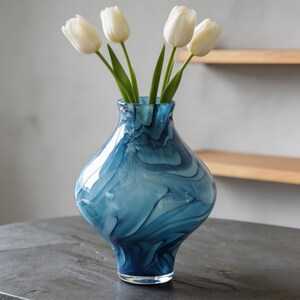 May include: A blue glass vase with a swirling pattern, holding five white tulips. The vase has a rounded shape and a clear base. The flowers have green stems and white petals. The vase is on a dark surface.