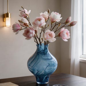 May include: A blue glass vase with a swirling pattern holds a bouquet of pink flowers. The vase has a rounded shape and a narrow neck. The flowers are in full bloom, with delicate pink petals and brown stems. The arrangement is on a dark wooden table.