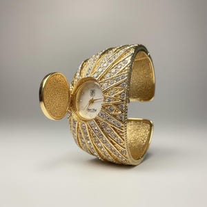 Könnte beinhalten: Eine goldfarbene Armbanduhr mit Klappdeckel. Das Zifferblatt ist weiß mit goldenen Zeigern und einem Logo. Das Armband ist mit klaren Kristallen besetzt. Ein auffälliges Schmuckstück.