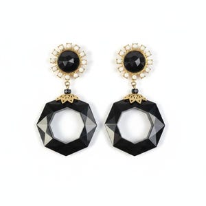 May include: A pair of statement earrings featuring a black geometric design. Each earring has a black center stone surrounded by white beads and gold-colored accents. The lower portion is a large, faceted, black hexagon.