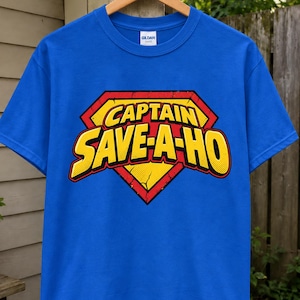 Puede incluir: Una camiseta azul brillante de manga corta con un estampado gráfico de un emblema de superhéroe amarillo y rojo. El emblema contiene el texto CAPTAIN SAVE-A-HO en letras amarillas negritas con un borde negro, sobre un fondo rojo desgastado.
