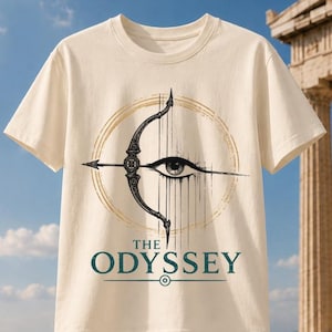 May include: A cream-coloured short-sleeve t-shirt featuring a graphic of a black bow and arrow centred within a gold circular design. Below the graphic, the text THE ODYSSEY is printed in dark green capital letters with a horizontal line.