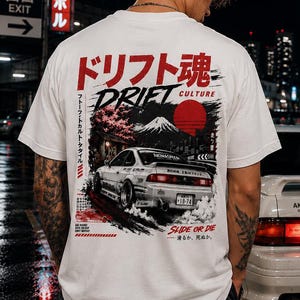 Puede incluir: Una camiseta blanca de manga corta con un estampado gráfico en la espalda. El diseño muestra un coche deportivo blanco, un sol rojo, una montaña y texto en japonés. El texto incluye 'DRIFT CULTURE' y 'SLIDE OR DIE' en tinta negra y roja.