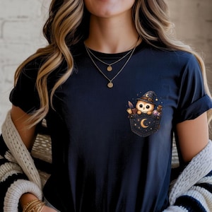 Può includere: Una t-shirt blu navy scuro con una stampa grafica di un piccolo gufo che indossa un cappello da mago all'interno di una finta tasca. Il design della tasca include una mezzaluna, stelle, una piuma e una boccetta di pozione.