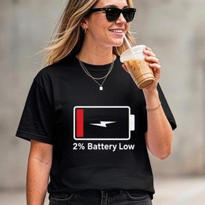Può includere: Una t-shirt nera a maniche corte con una grafica bianca raffigurante un'icona di batteria scarica con un segmento rosso e un fulmine bianco. Sotto l'icona, il testo 2% Battery Low è scritto in un carattere bianco senza grazie sul petto.