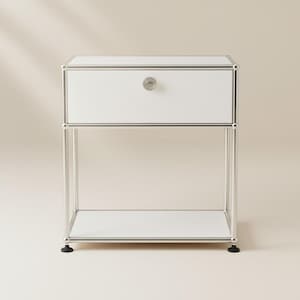 May include: A white bedside table with a single drawer and a lower shelf. The table has a silver metal frame and a round knob on the drawer. The table is supported by four legs and has black feet.