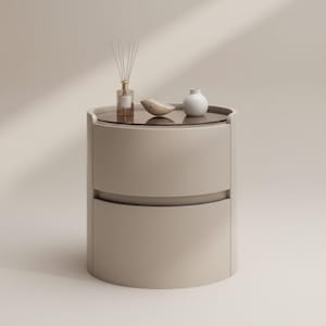 May include: A modern, beige bedside table with a round design. The top surface is a dark glass, holding a reed diffuser, a small white vase, and a decorative wooden bird. The table has two drawers and a minimalist aesthetic.