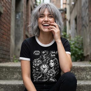 May include: Black ringer t-shirt with a white collar and sleeve trim. The graphic features a tiger, snake, woman, skull, and clock design in black and white. The shirt is worn by a person.