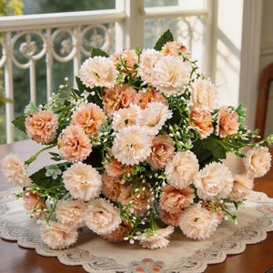 May include: A bouquet of artificial carnations in shades of peach and cream, with green foliage and small white accents. The flowers are arranged on a decorative, cream-colored doily, creating a soft and inviting floral display.