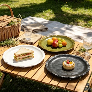 May include: Three round ceramic plates with wavy, irregular edges in white, olive green, and black. The white plate holds a sandwich, the green plate contains assorted fresh fruits, and the black plate features a small round cheesecake with berries.
