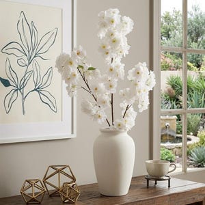 May include: A white ceramic vase holds a display of white cherry blossoms. The vase sits on a wooden table, alongside gold geometric sculptures and a teacup on a small black stand. A framed botanical print hangs on the wall.
