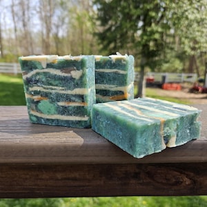 Puede incluir: Tres barras de jabón hechas a mano con un diseño en capas. El jabón presenta tonos de verde, blanco y naranja, creando un efecto veteado. Las barras son rectangulares y están sobre una superficie de madera, con un fondo exterior borroso.