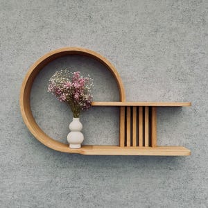May include: A modern, wooden wall shelf with a unique design. The shelf features a circular element and a rectangular section with vertical slats. A small white vase holds a bouquet of pink and white flowers, adding a touch of color.