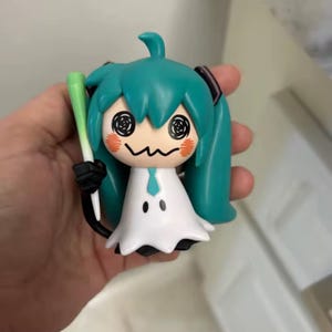 以下が含まれることがあります： ターコイズ色のツインテール、白いドレス、白黒の顔をしたキャラクターのフィギュア。フィギュアは緑と白のオブジェクトを持っています。キャラクターは頬にオレンジ色のチークマークがあります。