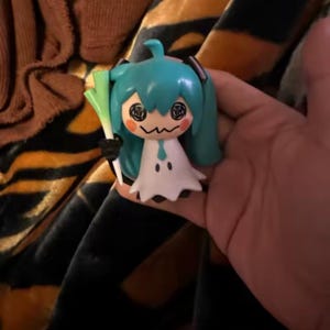 以下が含まれることがあります： ネギを持っている、水色の髪のキャラクターの小さなフィギュア。フィギュアは白いドレス、白黒の顔、小さな口をしています。フィギュアは人の手で持たれています。