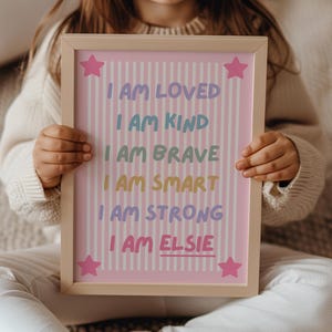 May include: A rectangular wooden frame containing a pink poster with white vertical stripes and four pink stars in the corners. The text reads: I AM LOVED, I AM KIND, I AM BRAVE, I AM SMART, I AM STRONG, I AM ELSIE, with each line in a different colour.