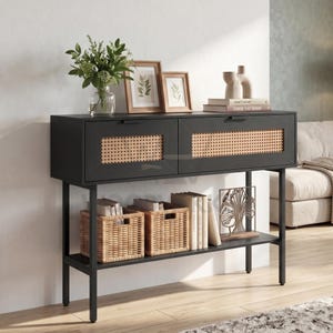 May include: A black console table with two drawers featuring natural woven cane panels and thin metal legs. The top surface holds a glass vase with green foliage, two framed botanical prints, and stacked books. Two woven baskets sit below.