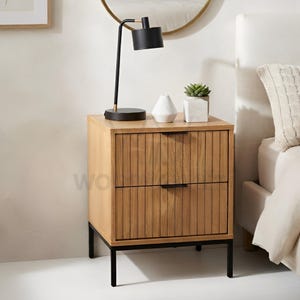 May include: A light oak bedside table with two drawers featuring vertical slats and thin black metal handles. The table stands on a black metal frame and holds a black desk lamp, a white geometric vase, and a small potted succulent plant.