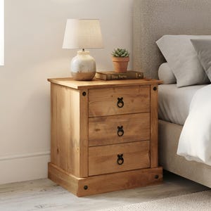 May include: A light brown wooden bedside cabinet featuring three horizontal drawers with black circular ring pulls. The piece has a rectangular top surface, a solid base, and visible wood grain patterns throughout the natural finish of the furniture.
