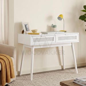 May include: A white desk featuring two drawers with cane webbing fronts and four tapered legs. The desk surface holds a yellow desk lamp, a small potted cactus, a picture frame, a bowl, and a closed laptop. The desk has a clean, rectangular design.