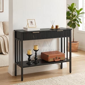 May include: A black console table with a fluted drawer front, two gold-toned handles, and a lower shelf. The top surface holds a framed picture and a reed diffuser, while the lower shelf displays two black candle holders and a brown suitcase.