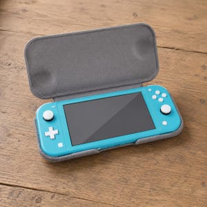 May include: A teal handheld gaming console with a gray screen, resting inside an open gray protective case. The console has white buttons and a black screen. The case is open, revealing the console on a wooden surface.